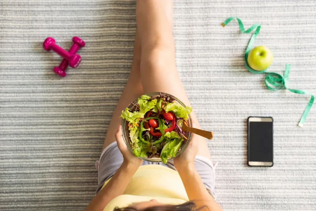 Como comer los dias de entrenamiento y ejercicio - Mujer con bowl de ensalada, movil y mancuernas